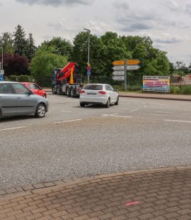 Die Aufnahme zeigt den Kreuzungsbereich an der Albert-Vater-Straße im Bereich der Abfahrt des Magdeburger Rings (MD-Ring). Im Vordergrund erstreckt sich eine mehrspurige Fahrbahn mit mehreren Fahrzeugen im fließenden Verkehr, darunter Pkw und ein Lkw mit Ladekran. Die Fahrbahn wirkt breit und stark frequentiert. Am rechten Fahrbahnrand befinden sich mehrere Wegweiser, die auf verschiedene Ziele im Stadtgebiet hinweisen. Dahinter ist eine eingezäunte Fläche mit Baustellen- bzw. Veranstaltungswerbung zu erkennen. Im Hintergrund prägen dichte Baum- und Grünflächen das Straßenbild. Links im Bild sind weitere Fahrspuren sowie die Zufahrt beziehungsweise Abfahrt des MD-Rings sichtbar. Der Himmel ist überwiegend bewölkt mit aufgelockerten Wolkenfeldern, wodurch eine leicht wechselhafte Wetterstimmung entsteht. Insgesamt vermittelt das Bild den Eindruck eines stark befahrenen Verkehrsknotens im urbanen Raum von Magdeburg.