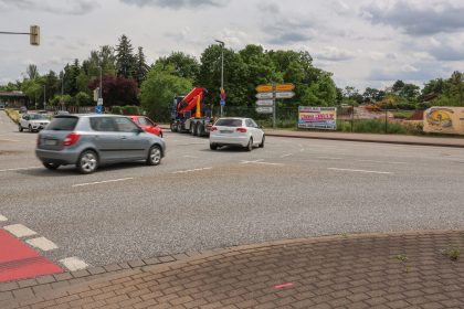 Die Aufnahme zeigt den Kreuzungsbereich an der Albert-Vater-Straße im Bereich der Abfahrt des Magdeburger Rings (MD-Ring). Im Vordergrund erstreckt sich eine mehrspurige Fahrbahn mit mehreren Fahrzeugen im fließenden Verkehr, darunter Pkw und ein Lkw mit Ladekran. Die Fahrbahn wirkt breit und stark frequentiert. Am rechten Fahrbahnrand befinden sich mehrere Wegweiser, die auf verschiedene Ziele im Stadtgebiet hinweisen. Dahinter ist eine eingezäunte Fläche mit Baustellen- bzw. Veranstaltungswerbung zu erkennen. Im Hintergrund prägen dichte Baum- und Grünflächen das Straßenbild. Links im Bild sind weitere Fahrspuren sowie die Zufahrt beziehungsweise Abfahrt des MD-Rings sichtbar. Der Himmel ist überwiegend bewölkt mit aufgelockerten Wolkenfeldern, wodurch eine leicht wechselhafte Wetterstimmung entsteht. Insgesamt vermittelt das Bild den Eindruck eines stark befahrenen Verkehrsknotens im urbanen Raum von Magdeburg.