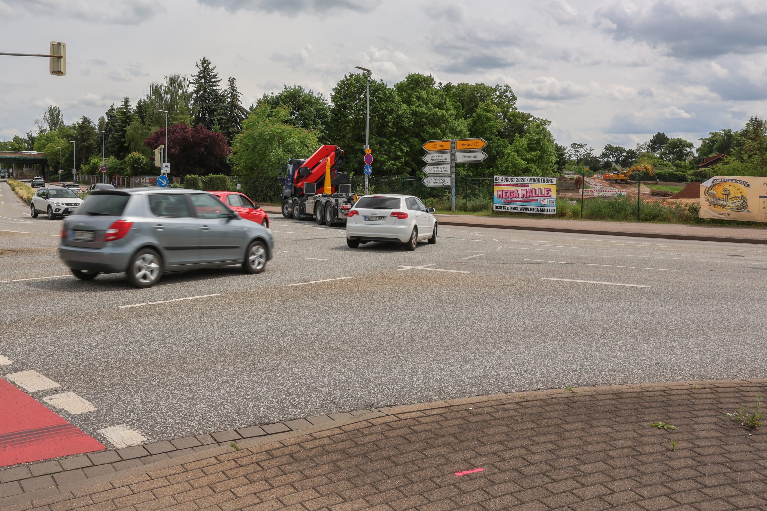 Die Aufnahme zeigt den Kreuzungsbereich an der Albert-Vater-Straße im Bereich der Abfahrt des Magdeburger Rings (MD-Ring). Im Vordergrund erstreckt sich eine mehrspurige Fahrbahn mit mehreren Fahrzeugen im fließenden Verkehr, darunter Pkw und ein Lkw mit Ladekran. Die Fahrbahn wirkt breit und stark frequentiert. Am rechten Fahrbahnrand befinden sich mehrere Wegweiser, die auf verschiedene Ziele im Stadtgebiet hinweisen. Dahinter ist eine eingezäunte Fläche mit Baustellen- bzw. Veranstaltungswerbung zu erkennen. Im Hintergrund prägen dichte Baum- und Grünflächen das Straßenbild. Links im Bild sind weitere Fahrspuren sowie die Zufahrt beziehungsweise Abfahrt des MD-Rings sichtbar. Der Himmel ist überwiegend bewölkt mit aufgelockerten Wolkenfeldern, wodurch eine leicht wechselhafte Wetterstimmung entsteht. Insgesamt vermittelt das Bild den Eindruck eines stark befahrenen Verkehrsknotens im urbanen Raum von Magdeburg.