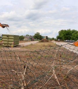Die Aufnahme zeigt ein geräumtes Baufeld in Blickrichtung Albert-Vater-Straße, mit Ausrichtung auf den Bereich An der Steinkuhle / Lorenzweg. Im Vordergrund befindet sich ein teilweise beschädigter Maschendrahtzaun, der das Baustellengelände von der Umgebung abgrenzt. Auf der Baustellenfläche sind umfangreiche Erd- und Räumarbeiten erkennbar. Links im Bild arbeitet ein orangefarbener Kettenbagger, der mit seinem Greifer beziehungsweise Löffel Material aufnimmt und bewegt. Daneben steht ein großer Absetzcontainer. Über das gesamte Baufeld verteilt liegen mehrere Haufen aus Holz-, Wurzel- und Grünschnittmaterial, die auf bereits erfolgte Rodungs- und Beräumungsmaßnahmen hinweisen. Im mittleren und hinteren Bereich der Fläche sind weitere Baumaschinen, Baustelleneinrichtungen sowie provisorische Fahrwege aus Schotter zu erkennen. Die Fläche selbst ist überwiegend unbefestigt und weist Spuren der Bauarbeiten auf. Entlang der rechten Seite begrenzen größere Baum- und Gehölzbestände das Baufeld. Im Hintergrund sind Gewerbe- und Betriebsgebäude sichtbar. Der Himmel ist überwiegend bewölkt, wodurch die Szene in einem diffusen Tageslicht erscheint. Insgesamt dokumentiert die Aufnahme den aktuellen Zustand eines vorbereiteten Baufeldes während laufender Erschließungs- und Beräumungsarbeiten im Bereich zwischen Albert-Vater-Straße, An der Steinkuhle und Lorenzweg.