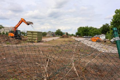 Die Aufnahme zeigt ein geräumtes Baufeld in Blickrichtung Albert-Vater-Straße, mit Ausrichtung auf den Bereich An der Steinkuhle / Lorenzweg. Im Vordergrund befindet sich ein teilweise beschädigter Maschendrahtzaun, der das Baustellengelände von der Umgebung abgrenzt. Auf der Baustellenfläche sind umfangreiche Erd- und Räumarbeiten erkennbar. Links im Bild arbeitet ein orangefarbener Kettenbagger, der mit seinem Greifer beziehungsweise Löffel Material aufnimmt und bewegt. Daneben steht ein großer Absetzcontainer. Über das gesamte Baufeld verteilt liegen mehrere Haufen aus Holz-, Wurzel- und Grünschnittmaterial, die auf bereits erfolgte Rodungs- und Beräumungsmaßnahmen hinweisen. Im mittleren und hinteren Bereich der Fläche sind weitere Baumaschinen, Baustelleneinrichtungen sowie provisorische Fahrwege aus Schotter zu erkennen. Die Fläche selbst ist überwiegend unbefestigt und weist Spuren der Bauarbeiten auf. Entlang der rechten Seite begrenzen größere Baum- und Gehölzbestände das Baufeld. Im Hintergrund sind Gewerbe- und Betriebsgebäude sichtbar. Der Himmel ist überwiegend bewölkt, wodurch die Szene in einem diffusen Tageslicht erscheint. Insgesamt dokumentiert die Aufnahme den aktuellen Zustand eines vorbereiteten Baufeldes während laufender Erschließungs- und Beräumungsarbeiten im Bereich zwischen Albert-Vater-Straße, An der Steinkuhle und Lorenzweg.